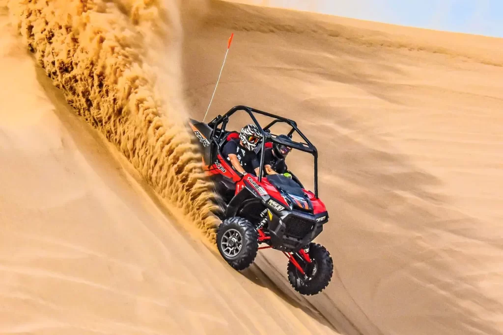 A vibrant orange dune buggy speeds across the golden sands of a vast desert in Dubai, with a clear blue sky overhead.